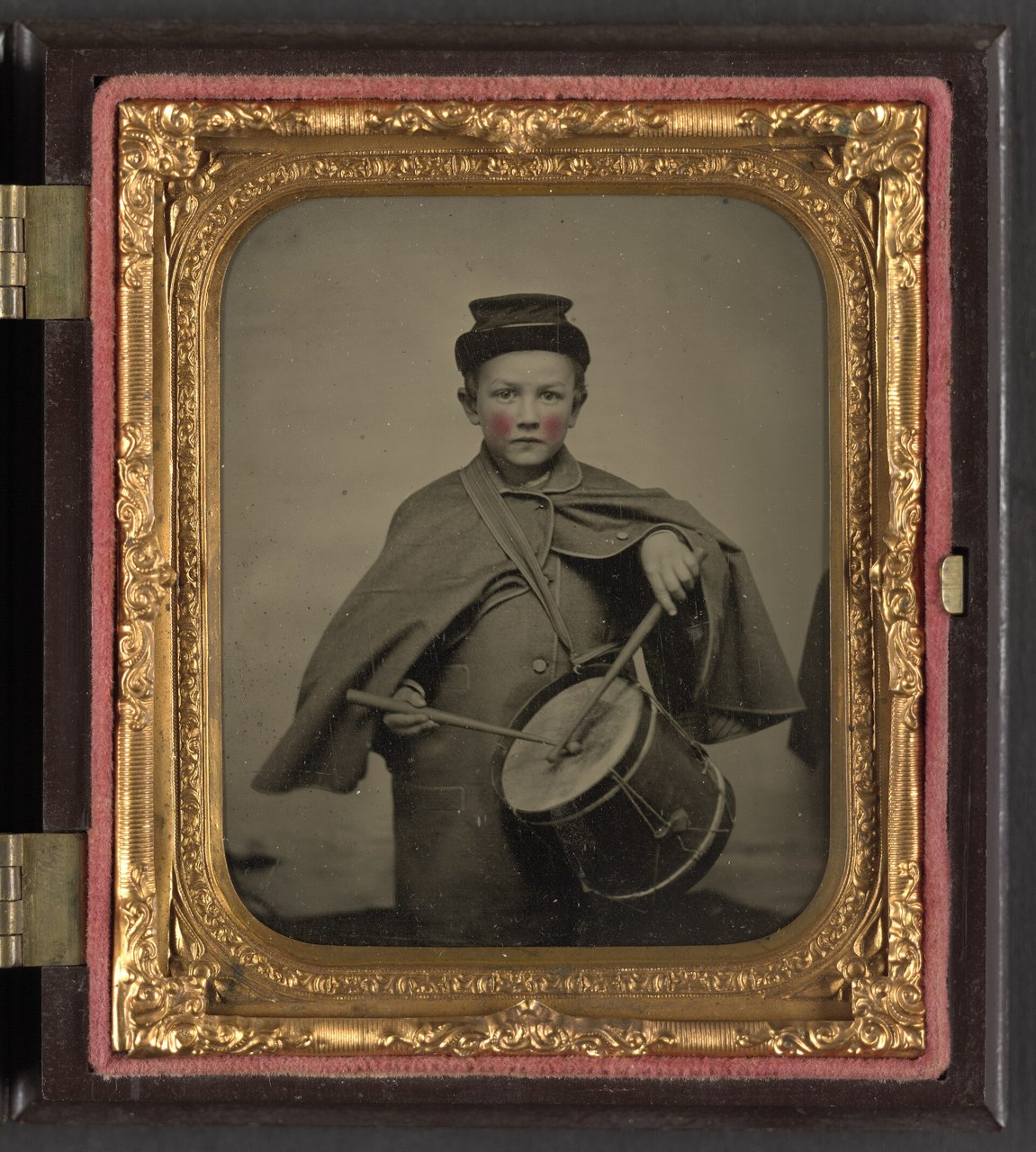 An old cased photograph in a gilt frame of a young boy in a soldier's cap and cape, holding drumsticks over a drum, his cheeks tinted pink