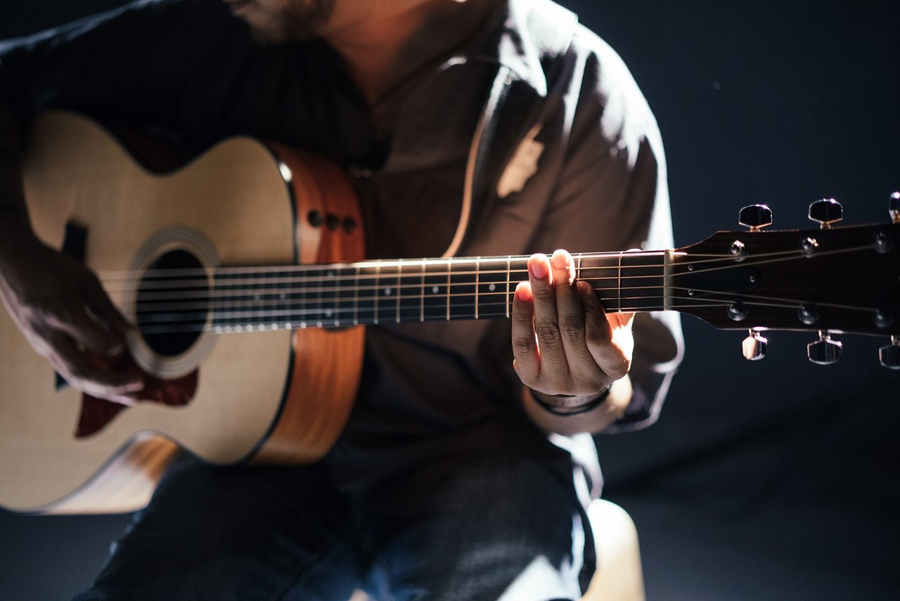 Close-up of a person's hands playing an acoustic guitar, one hand pressing the strings on the neck, lit by pale light against a dark background