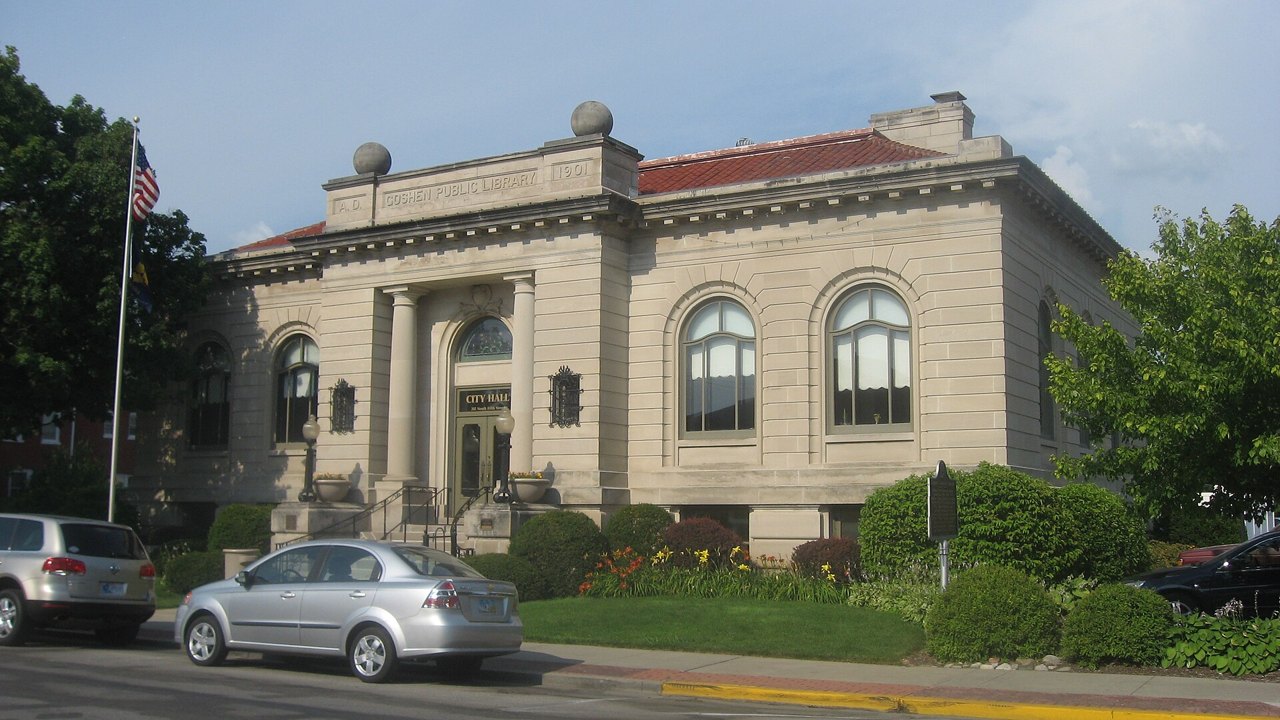 A small stone library building with a columned entrance, tall arched windows, and the words Goshen Public Library and the date 1901 carved along the top