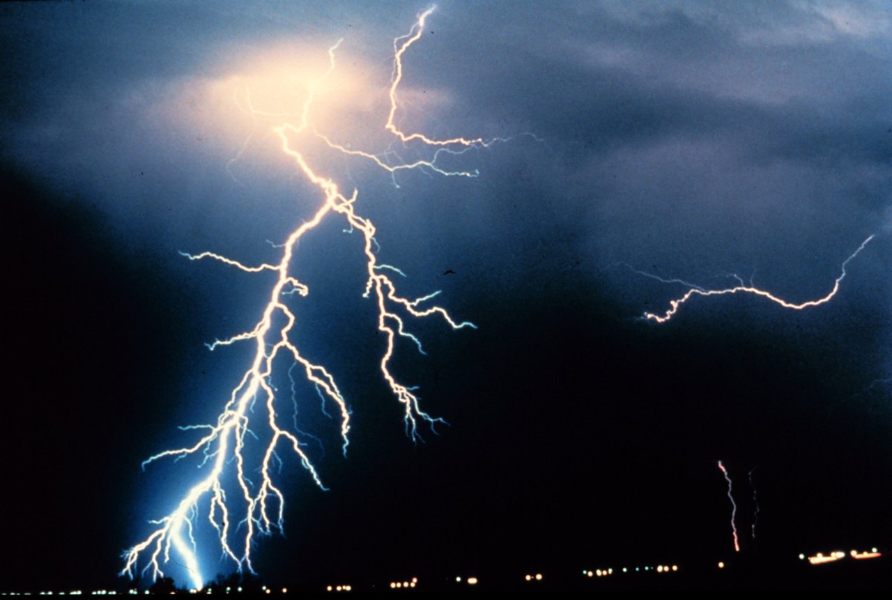 Bright forked lightning striking down from dark storm clouds at night, with a row of small lights along the ground far below