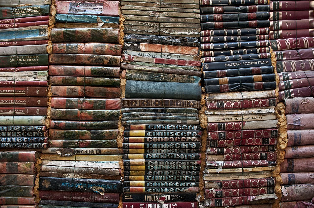 A wall built from tightly stacked old hardcover books, their worn spines in faded reds, greens, blues and browns, seen straight on