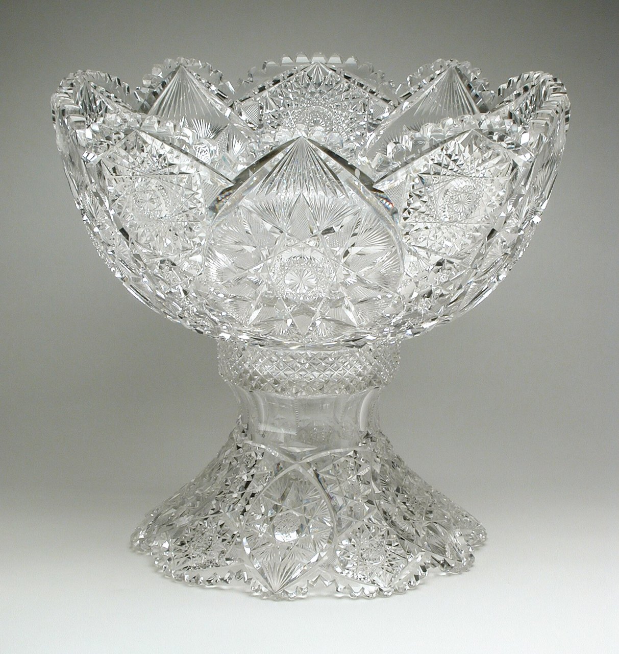 A large, heavily cut clear-glass punch bowl with a scalloped rim, standing on a matching cut-glass base, photographed against a plain gray background