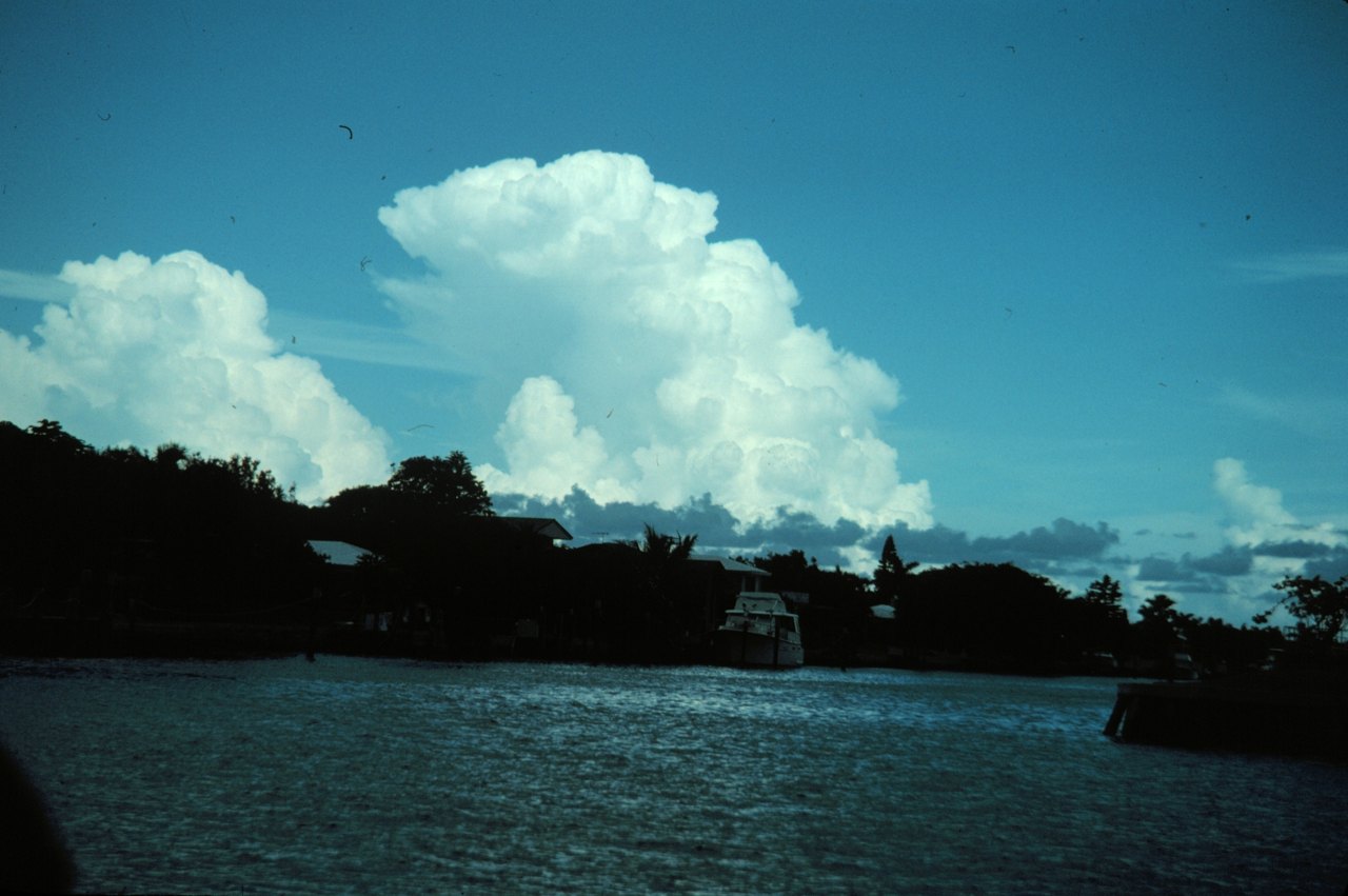 A huge, bright white thunderhead cloud towering into a deep blue sky above a dark line of trees and houses beside calm water, with a small boat moored at the shore
