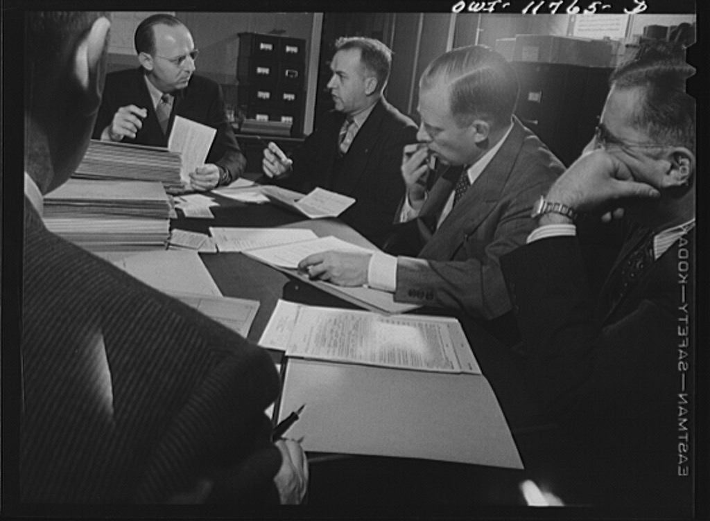 A black-and-white photograph of five men in suits seated around a long table piled with stacks of paper forms and folders, with filing cabinets behind them