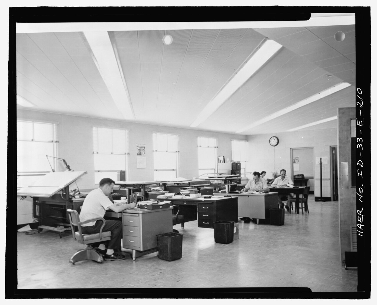 A black-and-white photograph of a bright office: two large tilted drawing tables stand along a wall of windows, a man in shirtsleeves works at a steel desk beside one of them, and more tables and desks recede toward a clock at the far end