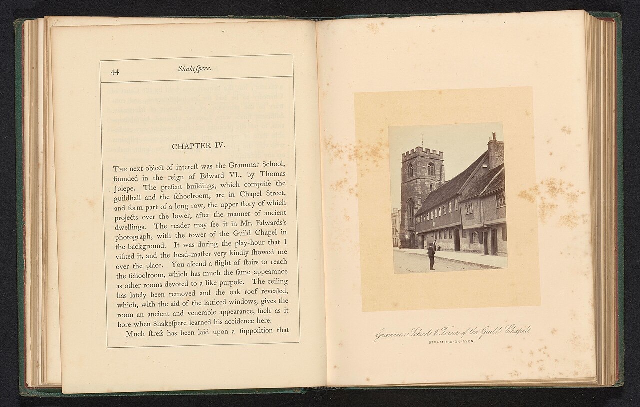 An old book lying open: printed text on the left page and, mounted on the right page, a pale albumen photograph of a long timber-framed building beside a stone church tower, with one man standing in the empty street