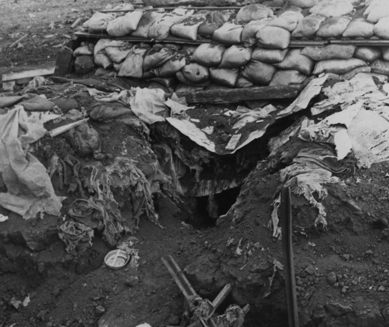 A black-and-white photograph of a bunker dug into bare earth, its low sandbag roof intact but its entrance torn open, with shredded cloth and scattered debris around the hole