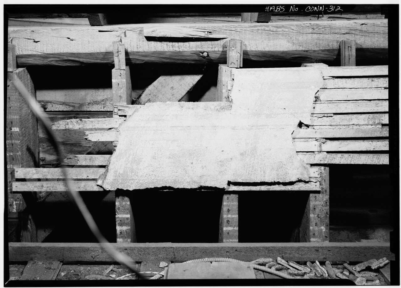 A black-and-white photograph looking down into an attic: thin wooden strips are nailed across the joists, and a broad slab of hardened plaster has squeezed through the gaps and set on top of them