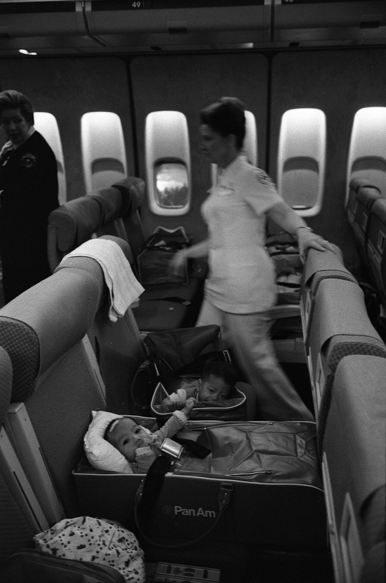 A black-and-white photograph inside an airliner cabin: two infants lie strapped into soft carry-cots set across a row of seats, while two uniformed flight attendants move along the aisle beside them