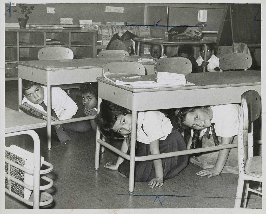 A black-and-white photograph of schoolchildren crouched on the floor underneath their classroom desks, several of them turning to look at the camera, with word cards and bookshelves along the wall behind