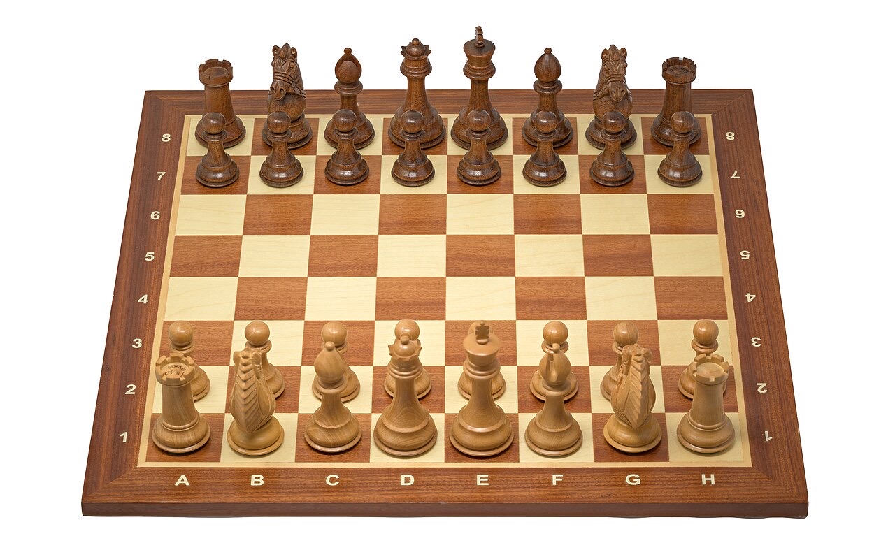 A wooden tournament chessboard with lettered and numbered edges, set up at the starting position with carved Staunton pieces in light boxwood and dark rosewood
