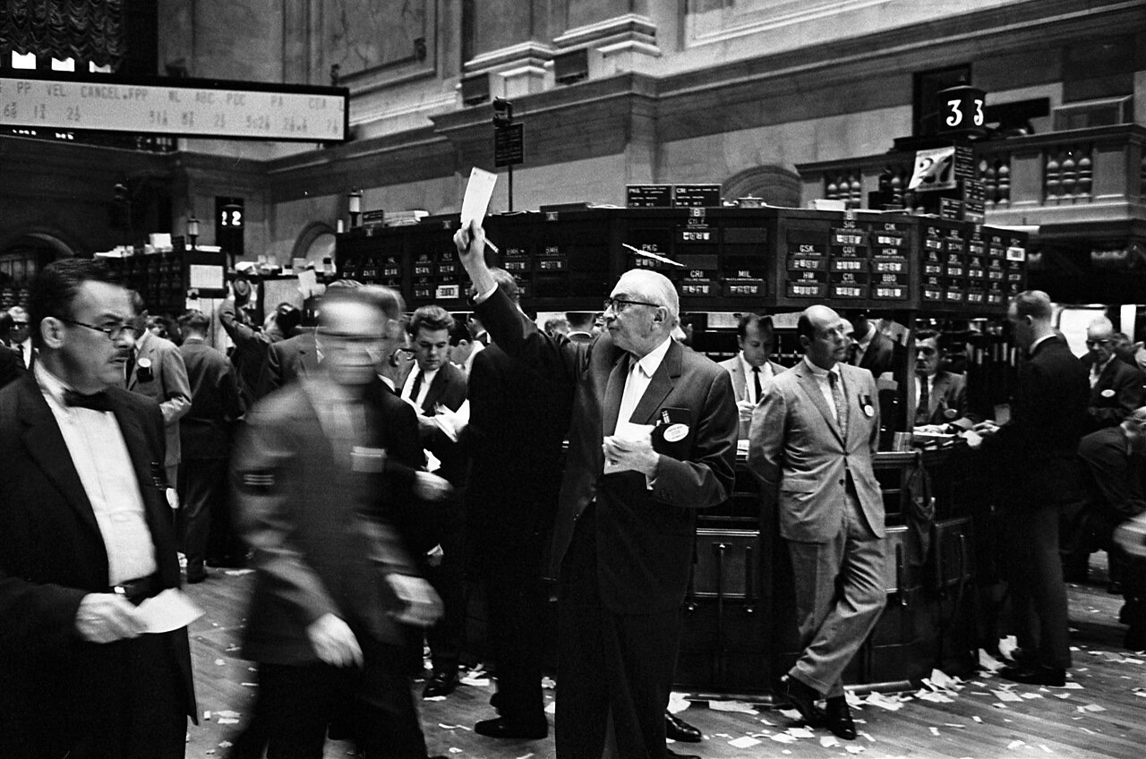 A black-and-white photograph of the New York Stock Exchange floor: suited brokers crowd around trading posts, one raises an order slip overhead, a quotation board glows above, and torn paper litters the floor