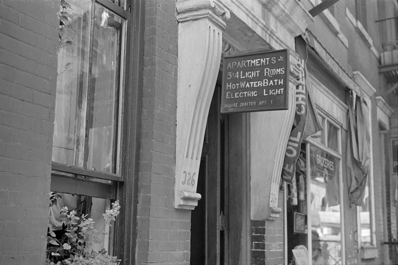 A black-and-white photograph of a brick apartment entrance with a hanging sign that reads Apartments, 3 and 4 light rooms, hot water bath, electric light, inquire janitor apt 1, beside a grocery window