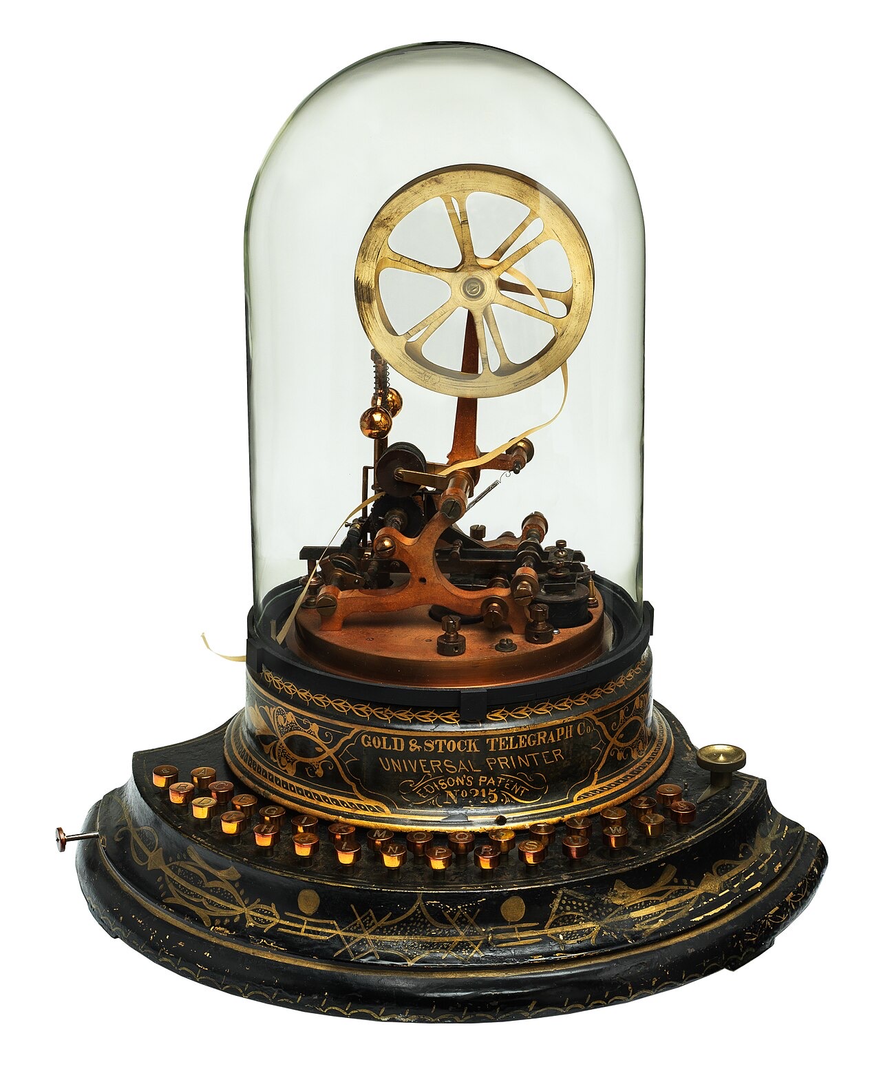A brass stock ticker under a tall glass dome on an ornate black and gold base: a spoked wheel feeds a narrow paper tape through the printing mechanism, above a ring of small round keys