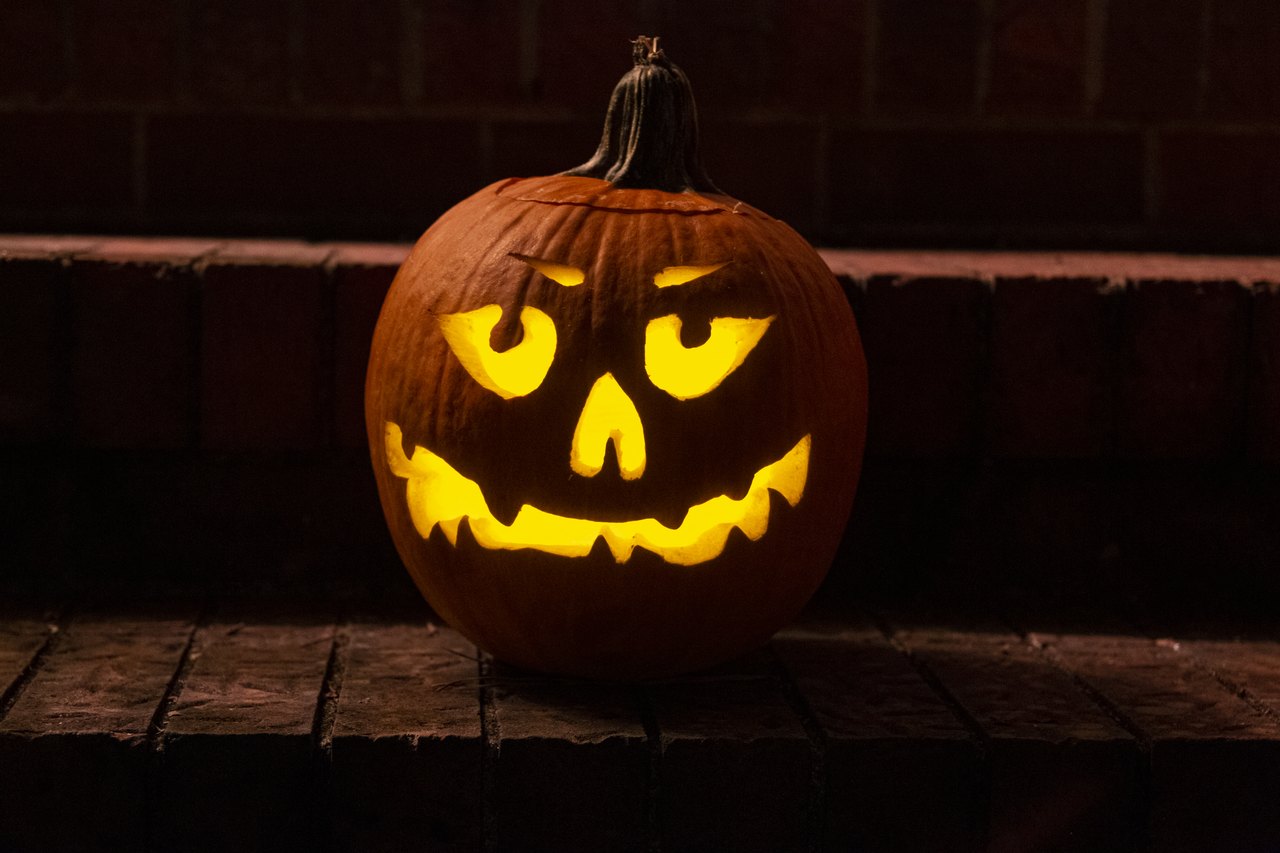 A carved pumpkin glowing from within on a wooden step at night, its triangular eyes and jagged grin lit orange against a dark brick wall