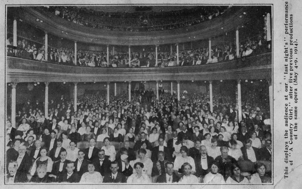 A packed theatre photographed from the stage in 1914, stalls and balcony filled with hundreds of seated people all facing the camera