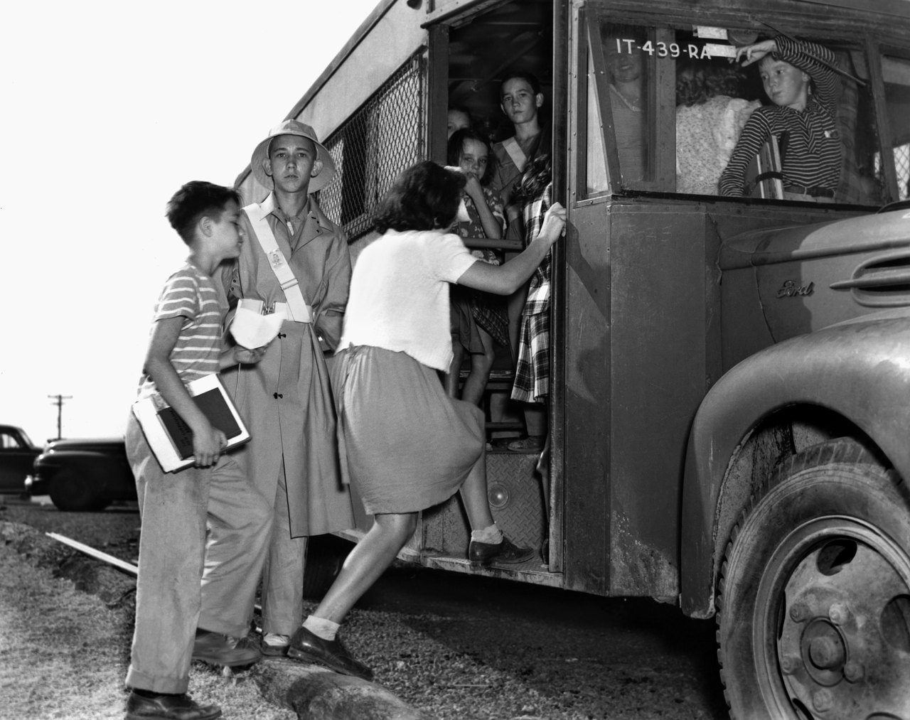 A 1944 black-and-white photograph of schoolchildren boarding a bus, one wearing a white safety-patrol belt across the chest and helping the others up the step