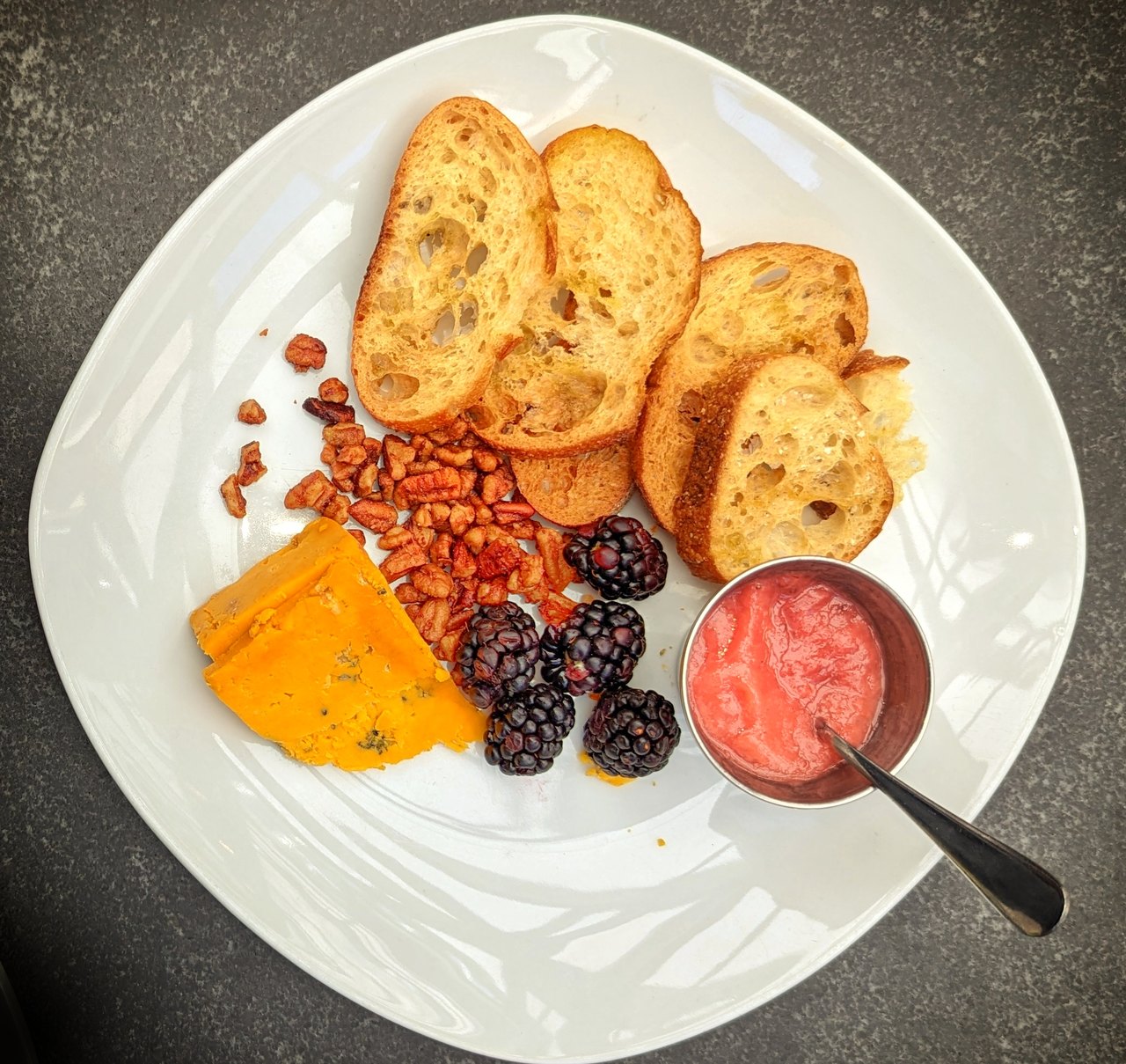 A white plate seen from above holding a wedge of orange blue-veined cheese, toasted bread slices, dried fruit, blackberries and a small pot of jam