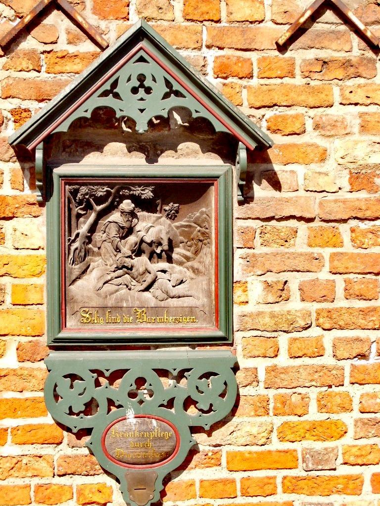 A small carved wooden alms box mounted on a brick wall under a pointed gabled roof, with a carved relief panel above the slot where coins are put in