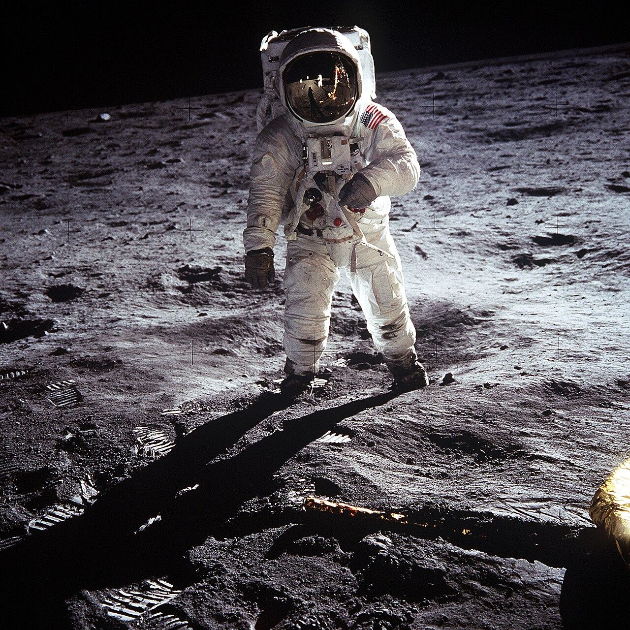 An astronaut in a white spacesuit standing on the gray surface of the Moon, his face completely hidden behind a curved gold visor that reflects the photographer and the lander