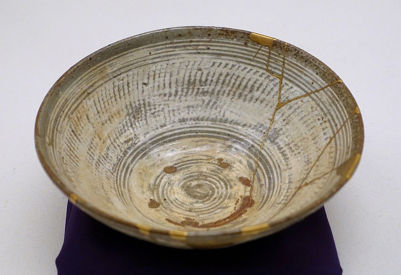 A shallow pale ceramic tea bowl seen from above, with several bright gold lines running across it where it was broken and joined again