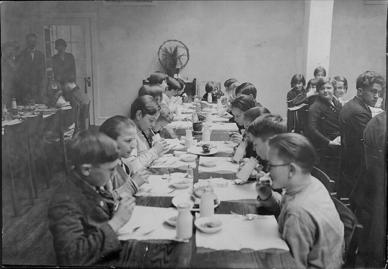 A black-and-white photograph of children seated along both sides of one long table in a school dining room, bowls and cups in front of them, adults standing at the back