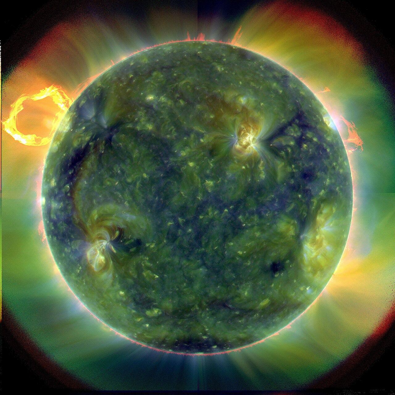 A full disk of the Sun photographed in ultraviolet light, its surface a mottled green and gold with a large loop of plasma arching off the left edge