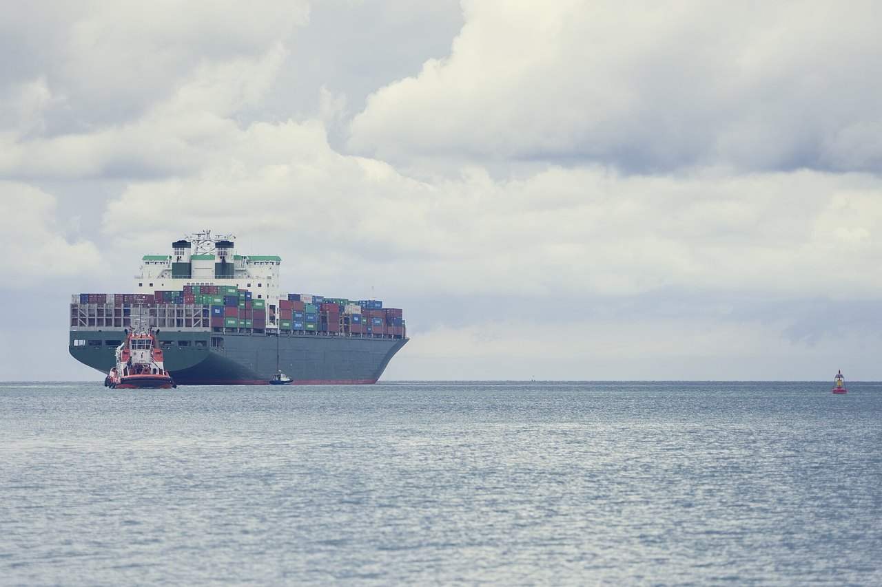 A large container ship stacked with colorful shipping containers moving across calm gray water under a cloudy sky, a small orange tugboat alongside
