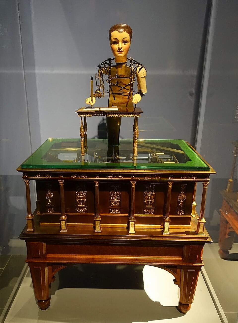 A museum automaton shaped like a boy, holding a pen over a small desk, mounted on an ornate brass cabinet with a glass top showing the gears inside