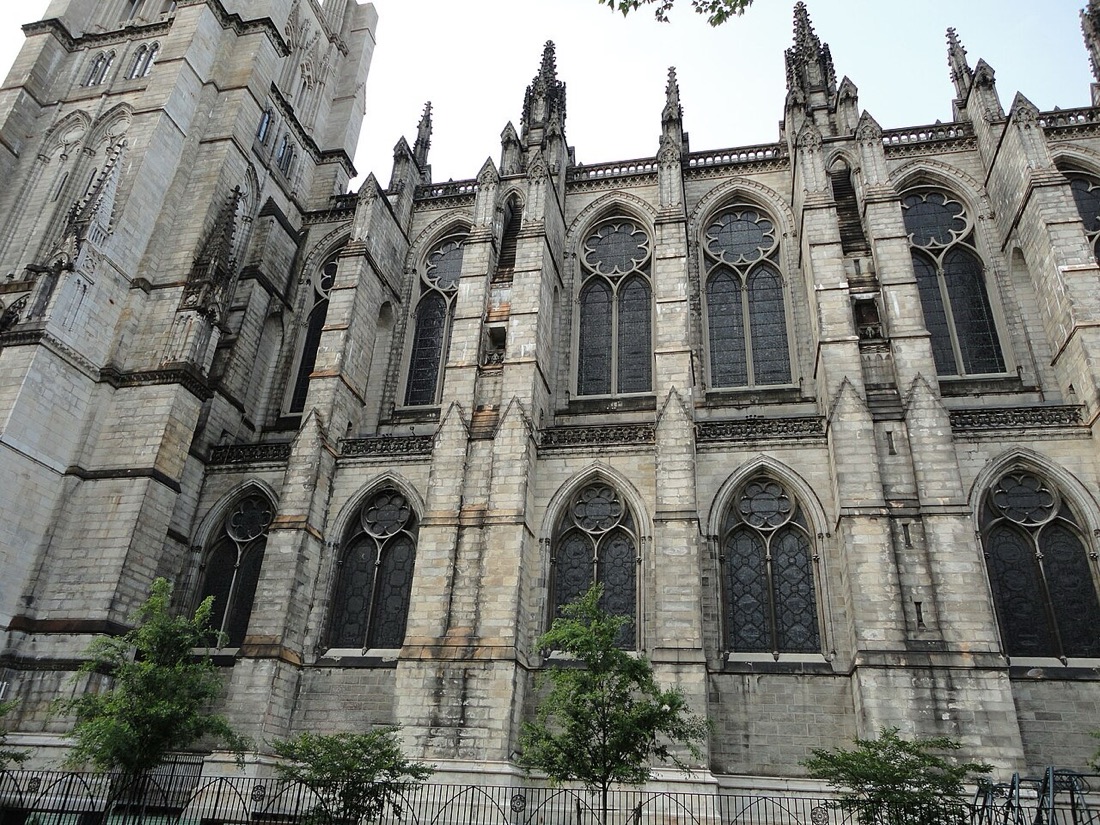 The gray stone side wall of a Gothic cathedral, with tall pointed windows, pinnacles and young trees in front