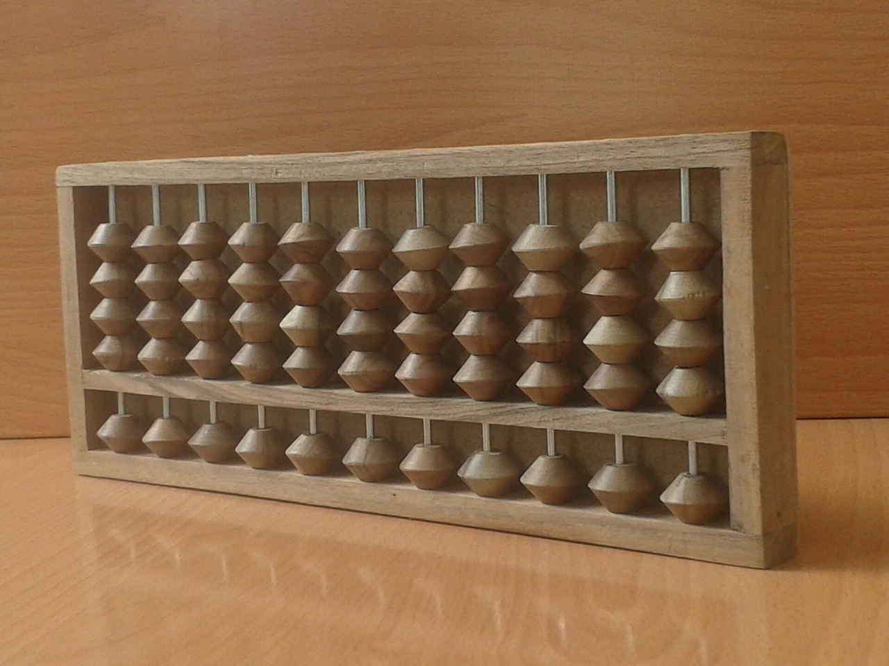 A wooden abacus standing on a wooden table, with rows of round beads on thin metal rods, four beads below a crossbar and one above on each rod