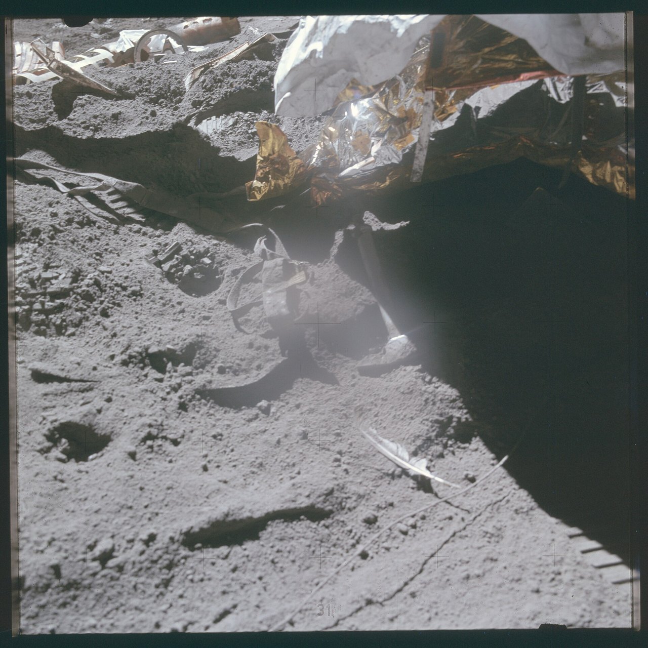 Gray, dusty lunar ground photographed from a spacecraft ladder: a small pale feather lies at lower right and a hammer lies near the center, with crumpled gold foil at the top