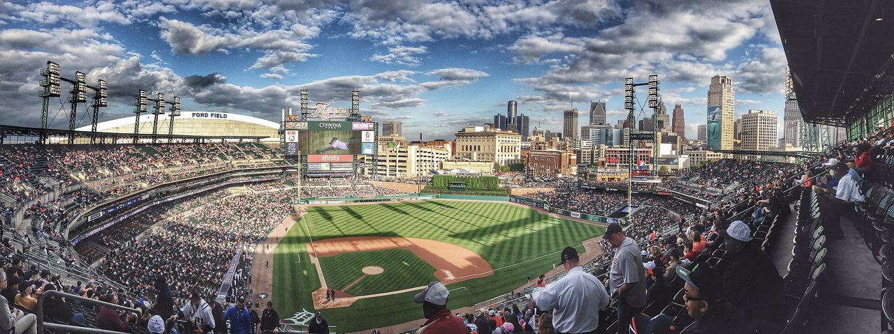 A wide view across a baseball stadium full of people on a sunny day, the green field below, city towers and a cloudy blue sky beyond