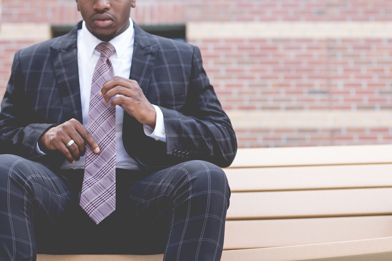 A man in a dark checked suit and white shirt sitting on a bench in front of a brick wall, both hands straightening his patterned necktie, his face out of the frame