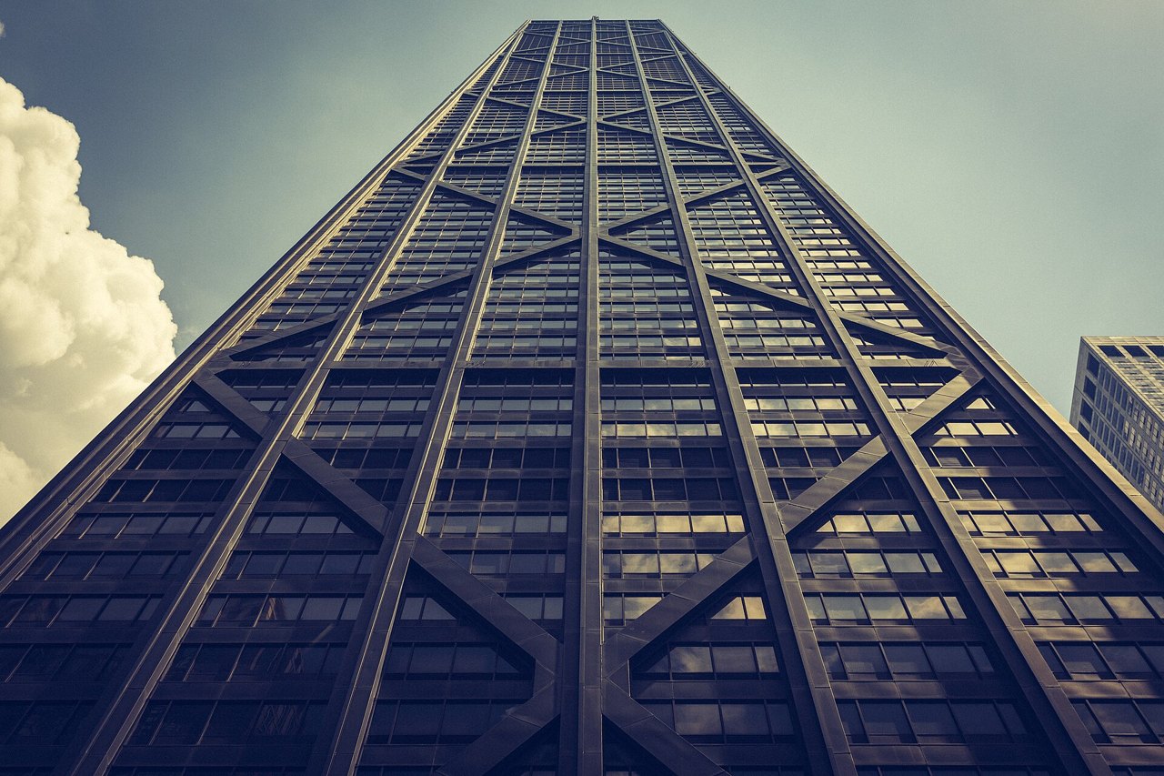 A very tall dark skyscraper photographed from the street looking straight up, its face crossed by huge X-shaped steel braces, against a pale sky with a white cloud