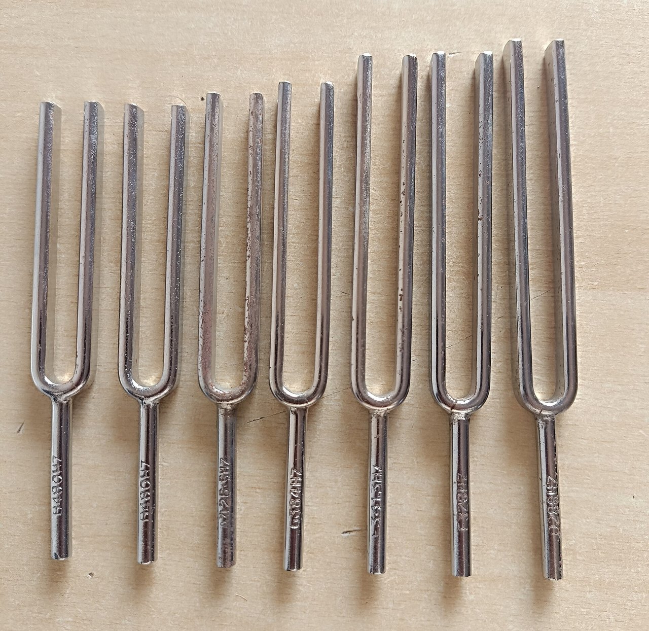 Seven steel tuning forks of slightly different lengths laid side by side on a pale wooden surface, each stamped with a number